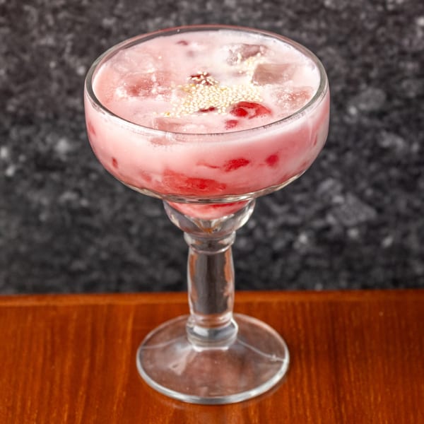 Pink Thai iced drink with red fruit pieces and white sesame seeds in a stemmed glass.