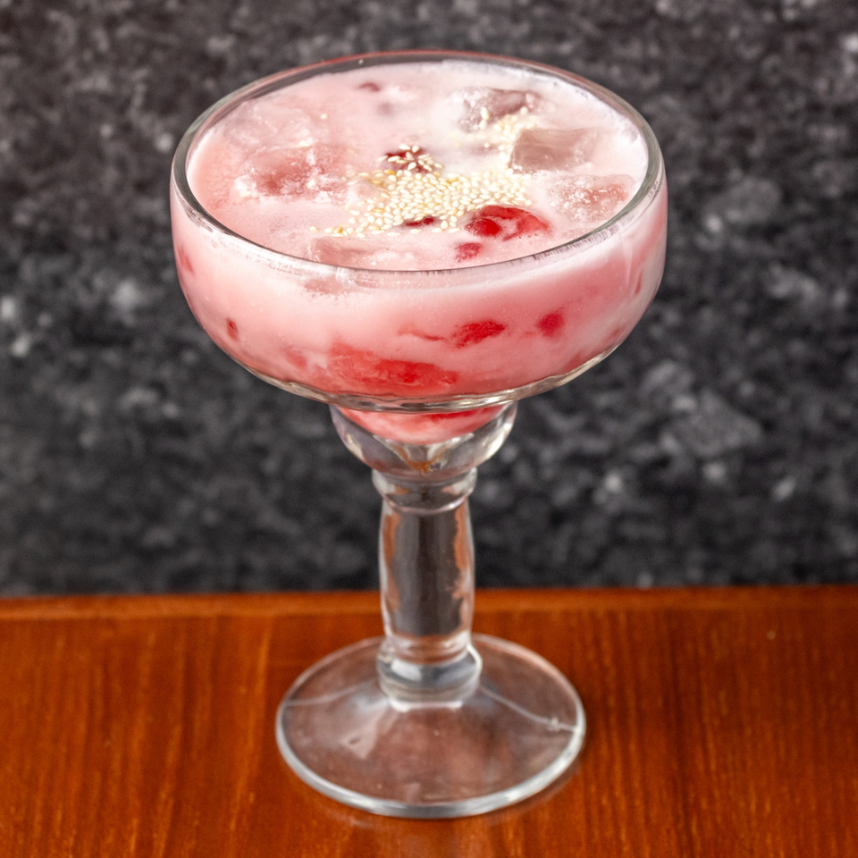 Pink Thai iced drink with red fruit pieces and white sesame seeds in a stemmed glass.