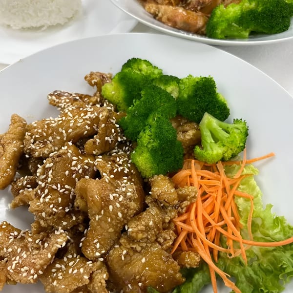 Sesame chicken with broccoli, shredded carrots, and lettuce on a white plate, with rice in the background.
