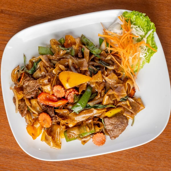 Thai Pad See Ew with beef, wide noodles, colorful vegetables, and a side of shredded carrots and lettuce.